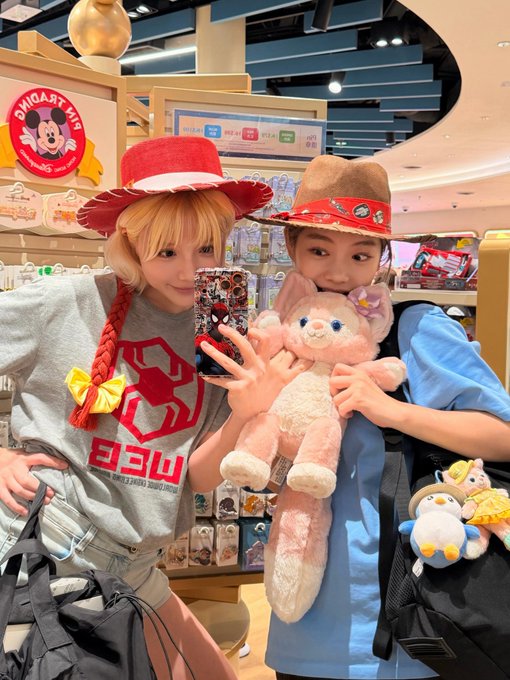Two women pose at a Disney store, one in a red hat with braided orange hair wearing a grey MFG shirt, the other in a brown cowboy hat holding a pink LinaBell plush toy.