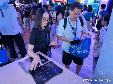 Two people at a busy expo or convention booth. A woman with glasses is pointing at a digital display on a counter, while a man next to her carries an ORCA branded tote bag. The website 'www.iorca.xyz' is visible in the bottom right corner.