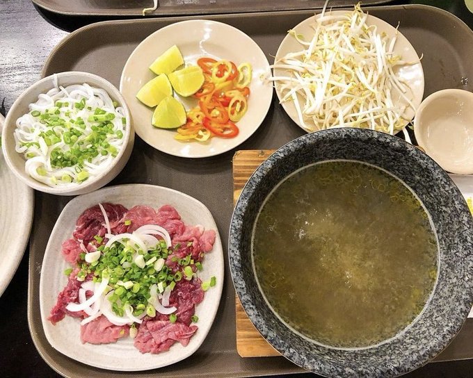 A tray of food featuring a bowl of clear broth, a plate of sliced beef with green onions and onions, a dish with sliced chili peppers and lemon/lime wedges, bean sprouts, and a bowl of rice noodles with green onions.