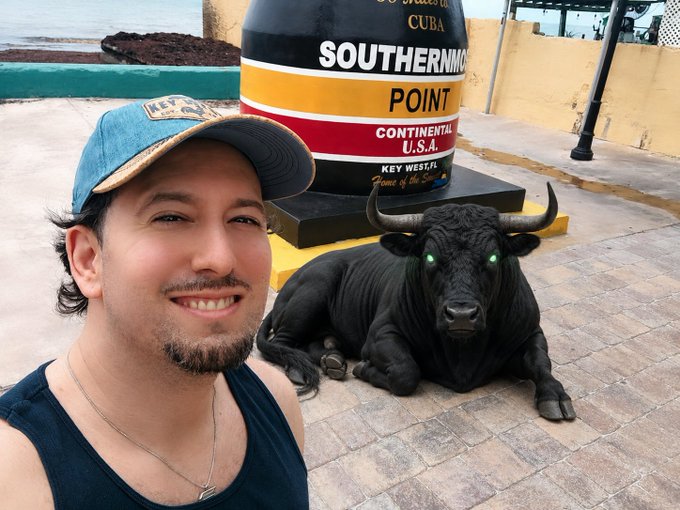 A man takes a selfie with a black bull statue at the Southernmost Point buoy in Key West, Florida, with the iconic "90 Miles to Cuba" landmark visible behind him as he poses with the mysterious glowing-eyed bovine referenced in his "$ANSEM" post.