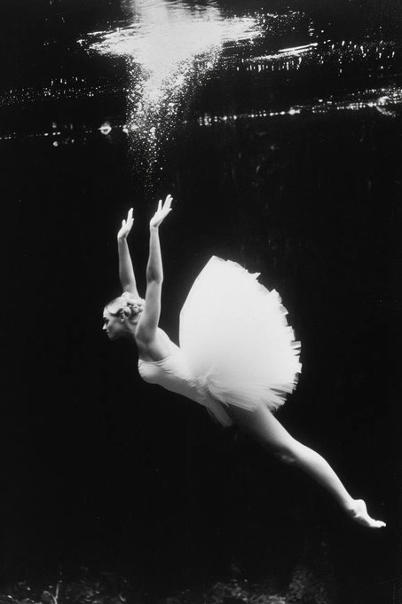 A black and white artistic photograph of a ballerina underwater, wearing a white tutu and reaching upward toward the surface.