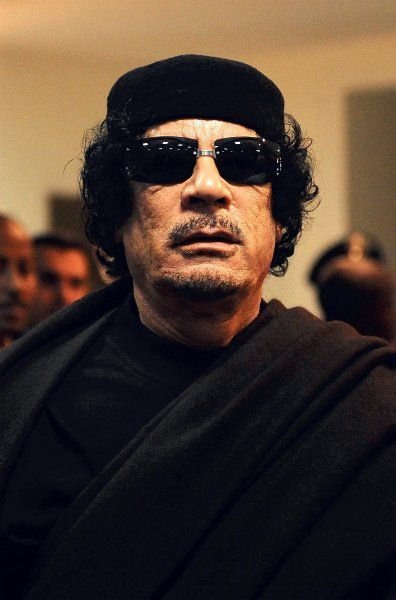 A man wearing dark sunglasses and a black cap, dressed in dark robes, with a serious expression, surrounded by other people in the background.