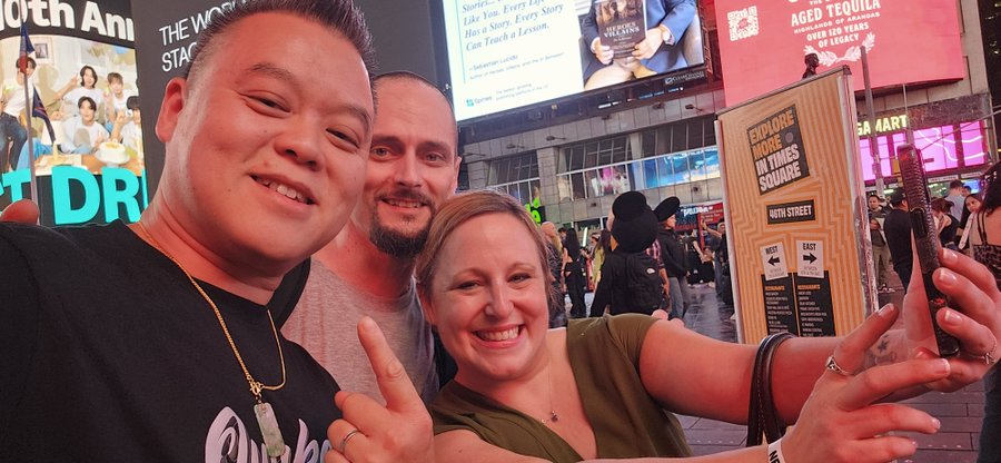 Three smiling friends pose for a selfie in Times Square, holding up peace signs, with the woman holding up a smartphone. Behind them, digital billboards advertise "Happy Vices" and "Aged Tequila Over 50 Years of Legacy," along with a sign reading "Explore More in Times Square: Ruth's Chris Steakhouse," capturing the lively NYC vibe of their #NFTNYC meetup.