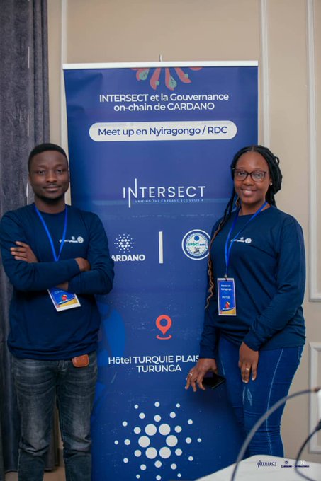 Two people in matching navy shirts with lanyards stand in front of a banner reading 'INTERSECT et la Gouvernance on-chain de CARDANO, Meet up en Nyiragongo / RDC' at Hôtel Turquoise Plaisir, Turunga.