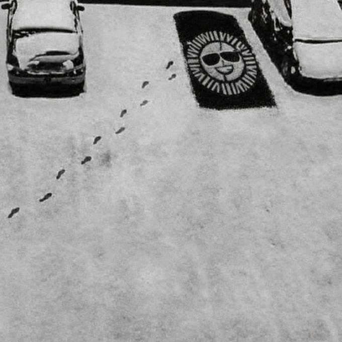 A grayscale image of a parking lot showing a trail of footprints leading across a snow-covered pavement toward a parking space marked with a rug featuring a smiling sun design, humorously tied to the caption "Parked my liquidity here" by visually representing a trader's "parking" of cash assets.