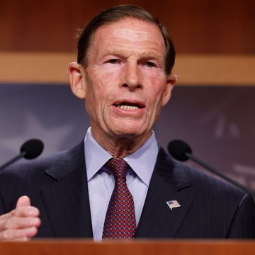 The image features a clean-shaven man in a dark suit with an American flag pin, speaking at a podium with microphones, identified by the accompanying post as US Senator Blumenthal discussing Tether's USDT cryptocurrency as a sanctions evasion tool for Iran.