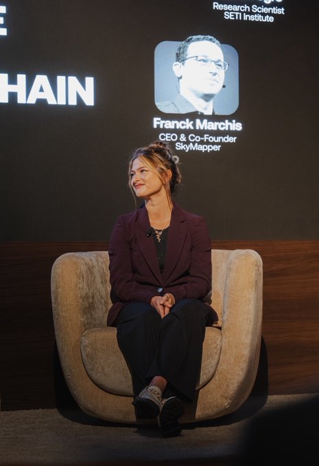 The image shows a woman with her hair pulled up, wearing a maroon blazer and dark pants, sitting in a beige armchair on a stage. Behind her on a screen is the text "Franck Marchis, CEO & Co-Founder, SkyMapper" along with a photo of a man and "Research Scientist, SETI Institute."