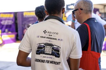 Two men stand in conversation, one wearing a white "Asi:one Your Personal AI" promotional t-shirt with "ASK US ONE" and "LOOKING FOR MERCH?" text, illustrating ASI:One's on-site presence as the official AI partner at the Goodwood Festival of Speed.