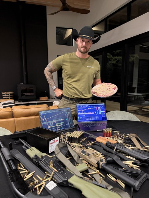 A man in a cowboy hat and olive green t-shirt holding a pizza, standing behind a table covered with various rifles, ammunition, and laptops displaying trading charts.