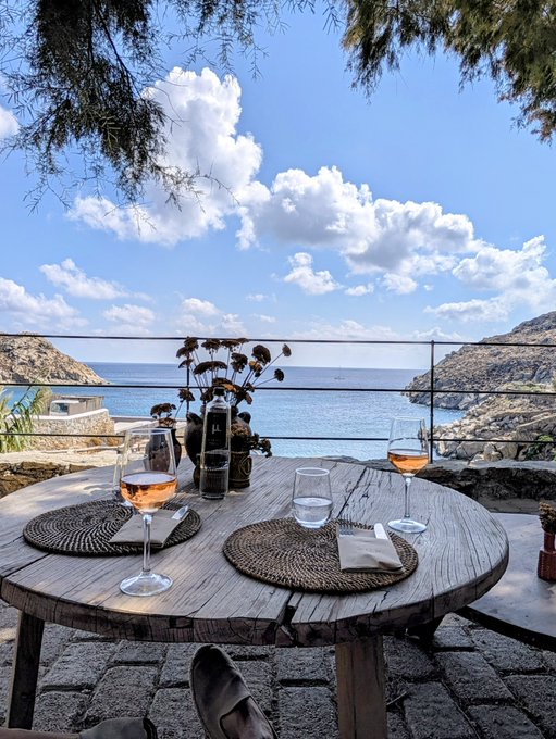 A rustic wooden table set with wine glasses and a meal overlooks a serene Mediterranean cove with dramatic cliffs and turquoise waters, evoking a tranquil escape that contrasts with the post's urgent call to pay attention to Bitcoin's 42% surge and the U.S. 10-year yield breaking out of a 3-year range. The juxtaposition suggests finding clarity away from markets, yet urges vigilance as significant financial shifts unfold.