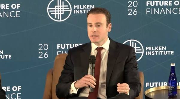 A man in a dark suit and red tie speaks into a microphone at the Milken Institute 'Future of Finance' 2026 panel, with a blue water bottle beside him.