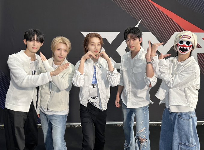 Five members of the K-pop group Stray Kids pose together in white outfits and jeans in front of a branded backdrop with their logo.