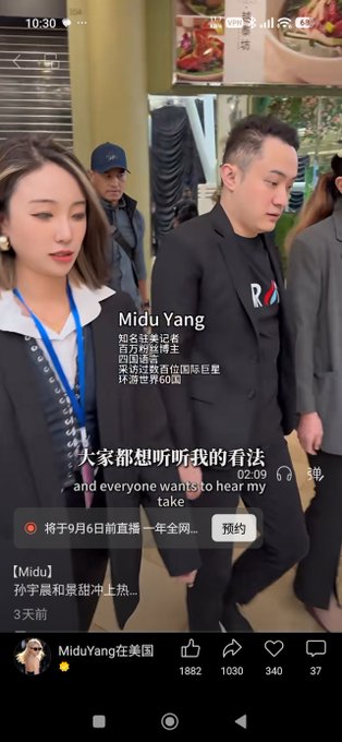 The image shows a video screenshot featuring Justin Sun walking with a woman, with an overlay of Chinese text from a reposted video by "MiduYang" that mentions famous journalist Bai Yansong and Justin Sun's international fans, with 500,000 views in the world. The caption "大家都想听听我的看法" ("and everyone wants to hear my take") reflects the post's interpretation that Sun is performing for media attention, as he appears focused and engaged while being filmed.