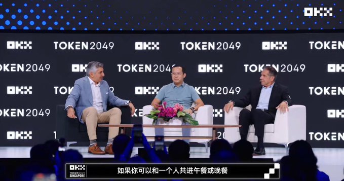 Three men seated in white armchairs on stage at the 'TOKEN2049' conference in Singapore, with the subtitle '如果你可以和一个人共进午餐或晚餐' visible at the bottom.