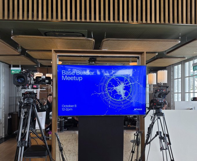 A large screen at a 'Base Builder Meetup' on October 8, 12-2pm, displays the event title next to the Base logo, with professional video cameras set up on tripods in the foreground.