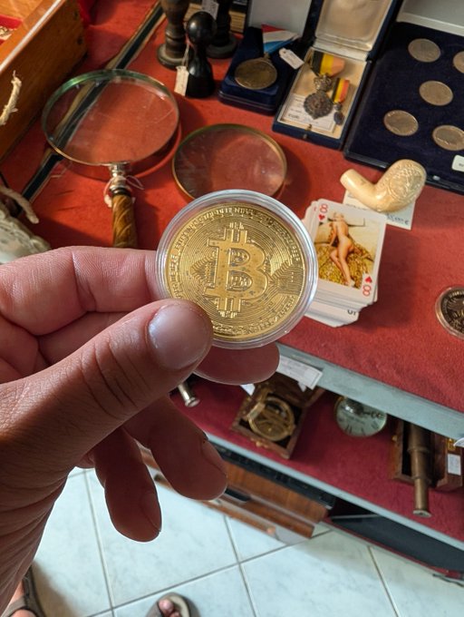 A hand holds up a gold Bitcoin commemorative coin with the "B" symbol and "BITCOIN" text visible, found among garage sale items like a magnifying glass, playing cards, medals, and various curiosities. The post humorously misrepresents this novelty collectible as the actual cryptocurrency, claiming a great "deal" of 500 € on a digital asset that is now worth far more, highlighting a common garage sale mix-up.
