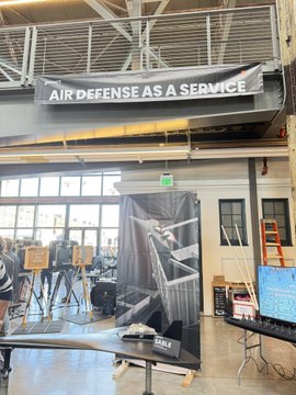 A banner reading "AIR DEFENSE AS A SERVICE" hangs above a booth at what appears to be Y Combinator's Demo Day, highlighting the growing presence of defense tech startups in the accelerator's lineup and reinforcing the post's point about its increasingly American defense-tech orientation.