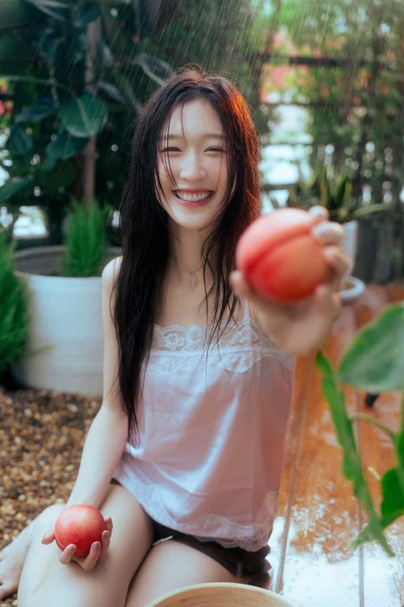 A smiling young woman with long dark hair, wearing a white lace camisole, sits outdoors in a garden setting holding peaches, with water streaming down around her.