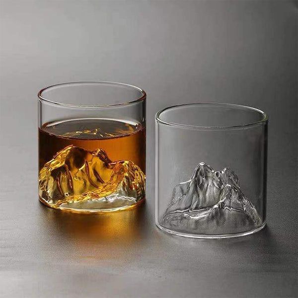Two glass tumblers featuring mountain-shaped designs at the base—one filled with amber liquid resembling whiskey, the other empty and clear.