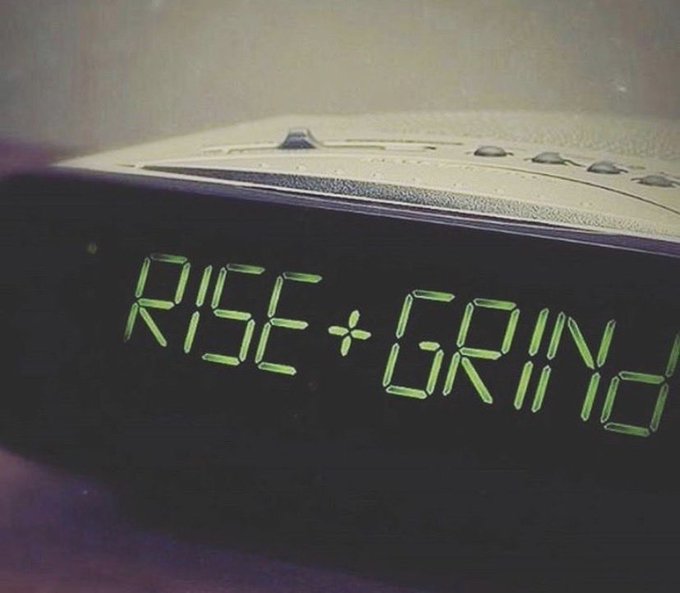 A close-up of a digital alarm clock displaying 'RISE + GRIN' in glowing green letters, styled like the 'Rise and Grind' motivational phrase.