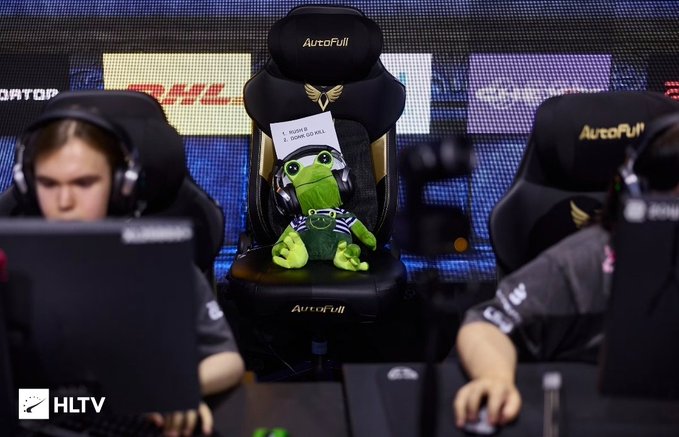 An esports gaming setup with players seated at AutoFull chairs, featuring a green Pepe the Frog plush toy holding a sign on the middle chair, with HLTV logo visible.