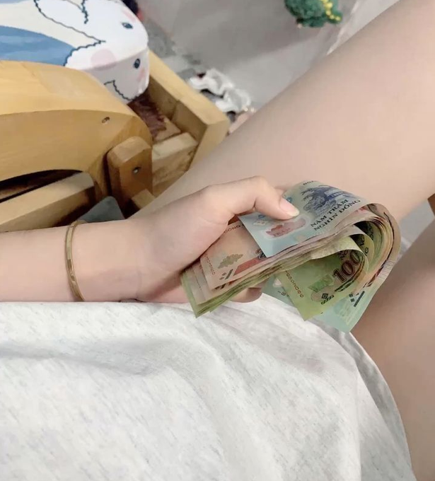 A hand holds a stack of mixed international banknotes including what appears to be a 100 denomination bill, with a gold bracelet on the wrist.