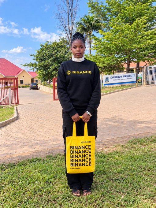 A woman stands outdoors in front of Kyambogo University holding a yellow Binance-branded tote bag that reads 'BINANCE BINANCE BINANCE BINANCE' while wearing a black Binance sweatshirt.