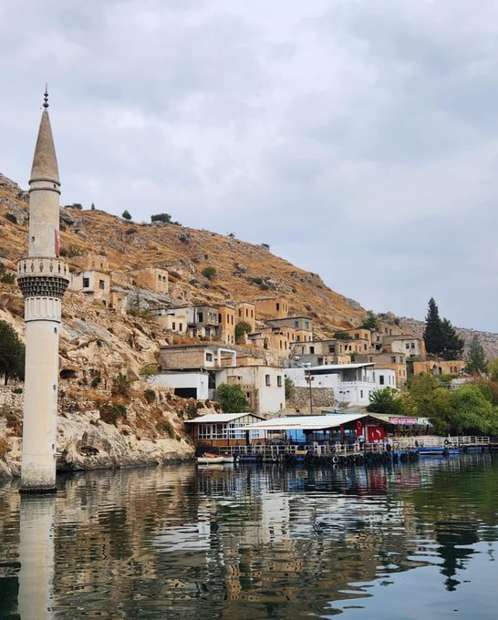 A scenic view of a village built into a rocky hillside along the water, featuring a tall minaret in the foreground and waterfront restaurants.