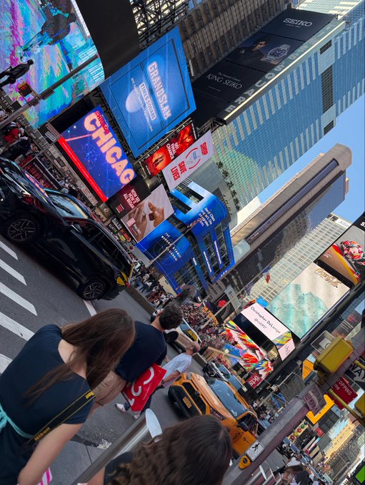 A street-level view of Times Square in New York City shows a crowded intersection with yellow taxis, billboards including 'CHICAGO' and 'SEIKO', and pedestrians.