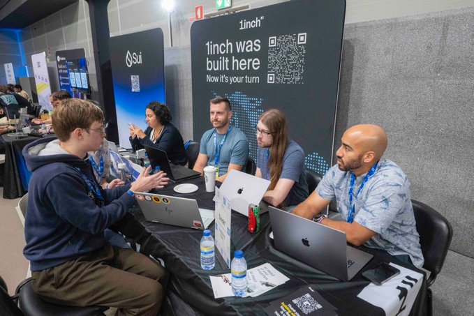 A group of people sit at a booth with laptops; banners behind them read "1inch was built here. Now it's your turn" with a QR code and a "Sui" logo.