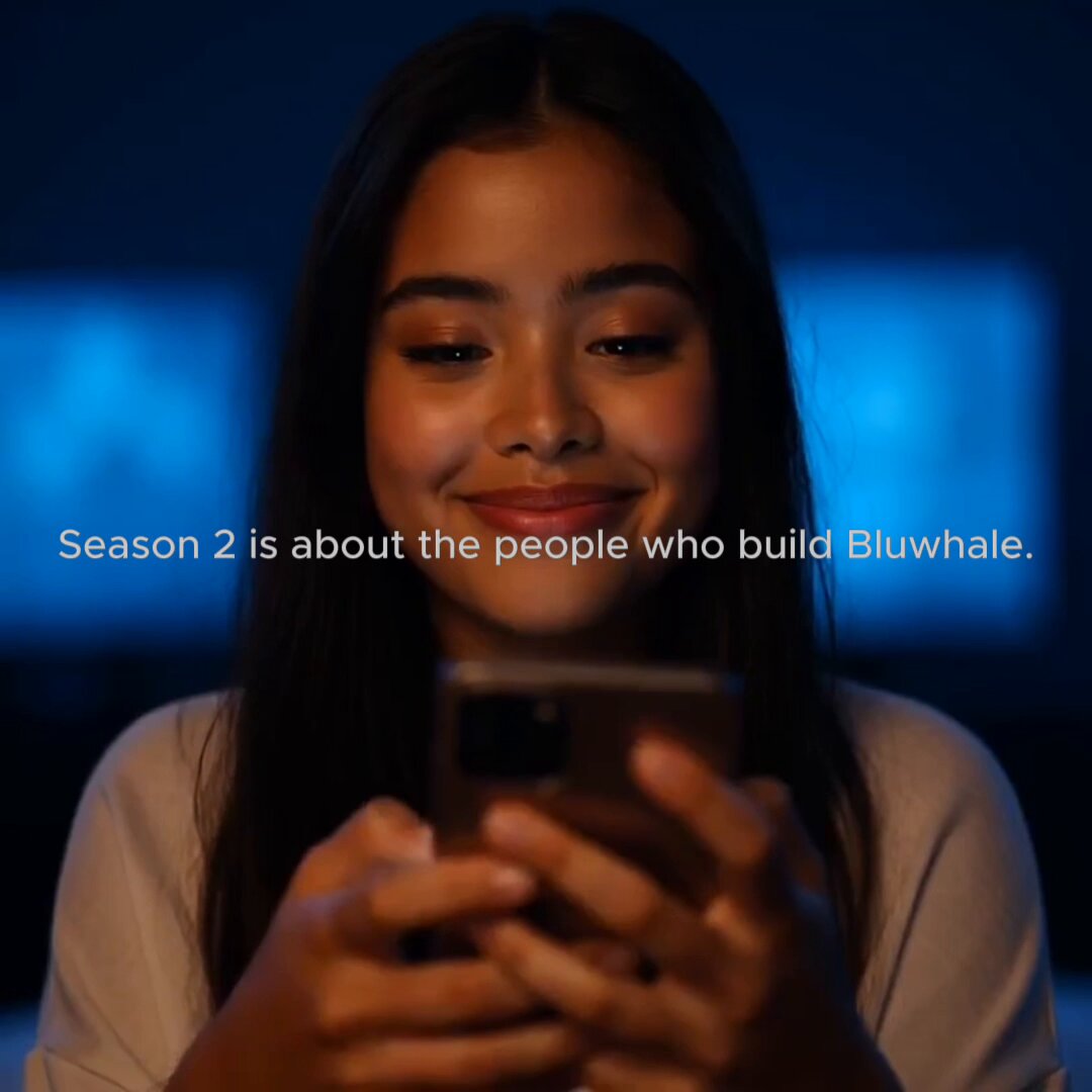 The image shows a woman holding a cell phone with her hands while looking at it. The caption for this image reads, "Season 2 is about the people who build Bluwhale. Season 2 is about the community that helps grow the Bluwhale ecosystem." This suggests that the image is related to an advertisement or promotional material for the Bluwhale project and highlights the importance of community engagement in supporting and growing the project.