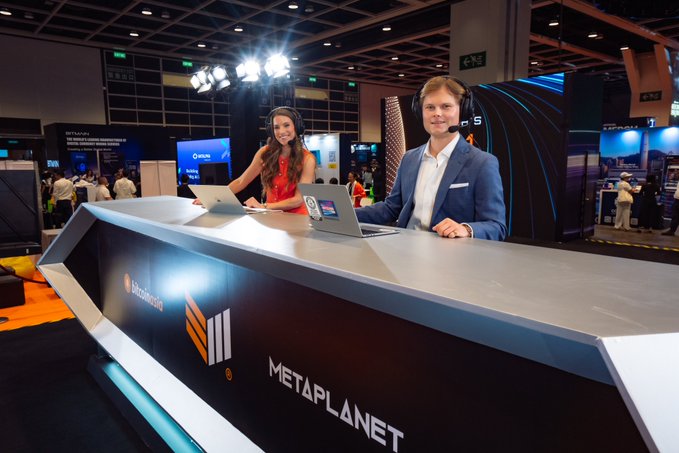 The image shows Bitcoin TV anchors seated at the "METAPLANET" branded live news desk at Bitcoin Conference Asia, wearing headsets while presenting from laptops, which ties into the post announcing BMTV anchor Grace Remington and market analyst Sean Hagan hosting coverage from the conference ahead of the network's September 14th launch.