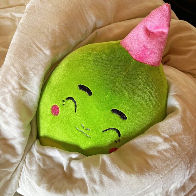 A green plush pillow with a cute smiling face and a pink horn or ear, resting in white bedding.