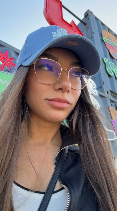 A young woman takes a confident selfie in front of a colorful mural featuring neon signs of food items like burgers and chili dogs, wearing a blue baseball cap, glasses, and a leather jacket. Her poised, unbothered expression perfectly captures the smug "look" referenced in the caption about flexing a $MAME portfolio.