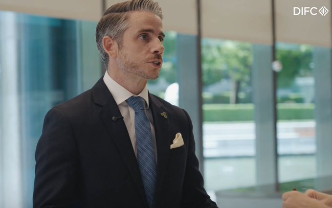 A man in a dark suit with a blue tie and yellow pocket square is being interviewed, with a 'DIFC' logo visible in the upper right corner of the frame.