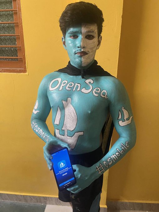 A young man with full-body turquoise and silver face paint featuring "OpenSea" branding, an anchor logo on his chest, and "#IOSMobile" written on his arms, holds a smartphone displaying an airdrop notification from OpenSea while waiting for the token drop.