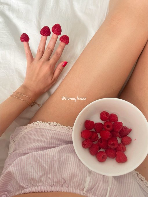 A hand adorned with raspberries on the fingertips reaches toward a bowl filled with fresh raspberries, resting beside legs in lace-trimmed pajamas on white sheets, with the watermark "@honeytoezz" visible. The playful "GM" (good morning) caption pairs perfectly with this sweet, berry-themed morning scene.