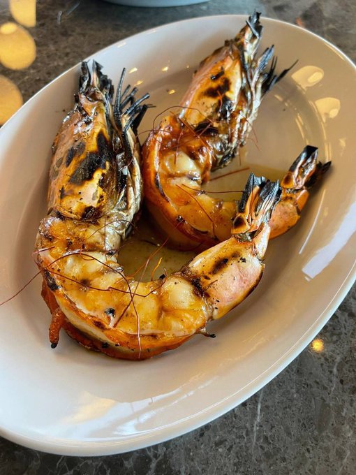 Three large grilled prawns are served on an oval white plate, showing characteristic grill marks and buttery glaze.
