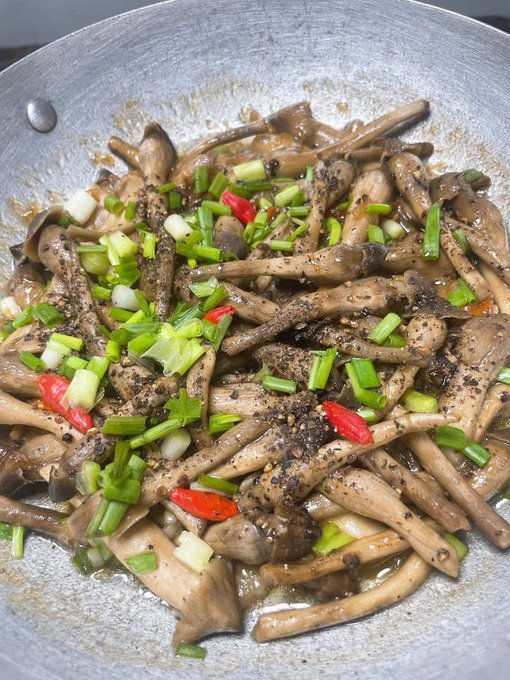 A sizzling pan of stir-fried mushrooms garnished with green onions, red chilies, and cracked black pepper, served in a rustic metal skillet. The Vietnamese caption invites followers to join "Zy" for lunch, playfully asking if anyone can guess what dish is pictured.