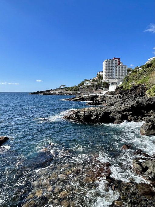 iced tea house with a red roof and white walls sits on a rocky cliff overlooking the ocean, with a clear blue sky above.