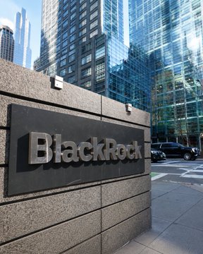 A "BlackRock" corporate sign is mounted on a stone wall outside a glass-fronted office building, with a parked SUV visible on the street. The image illustrates the subject of the accompanying article about BlackRock's head of digital assets advocating for Bitcoin as a hedge against mounting U.S. debt.