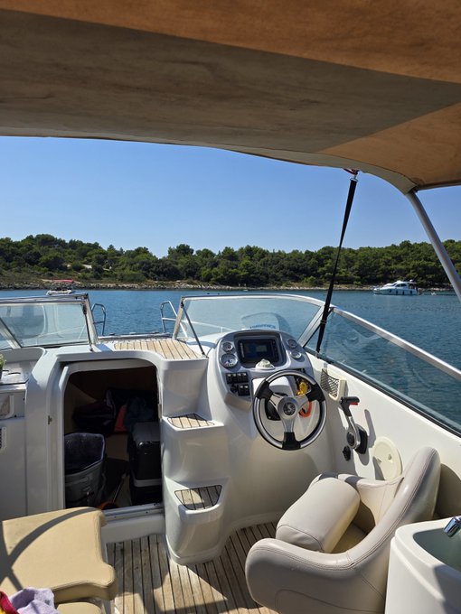 View from the helm of a motorboat docked in calm blue waters, with green tree-lined shores visible in the distance under a clear sky.