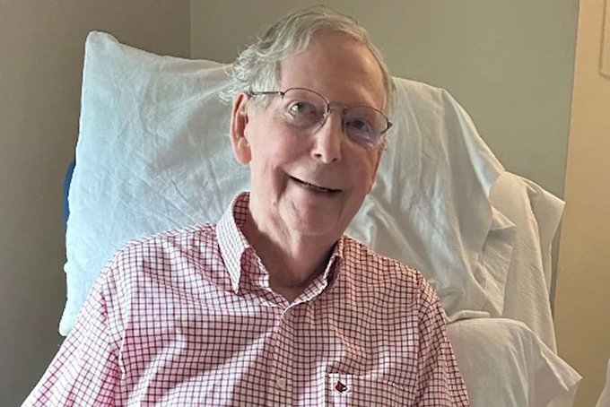 A smiling elderly man with gray hair and glasses, wearing a red and white checkered shirt, sits in a chair with a white pillow behind him in what appears to be a medical or care setting.