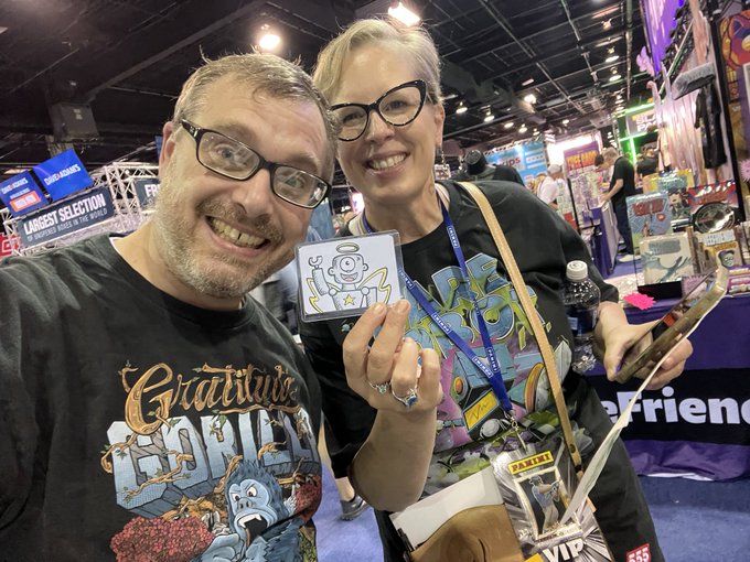 A man wearing a 'Gratitude Dead' Grateful Dead t-shirt and a woman wearing glasses pose for a photo at what appears to be a comic convention, with the woman holding a small sticker card.