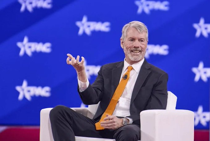 A gray-haired man in a dark suit and bright orange tie speaks animatedly while seated on a white chair at a CPAC event, gesturing with his right hand.