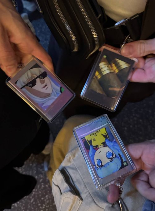Three hands display anime-style acrylic keychain charms featuring characters like Bartz from Final Fantasy V and a crying figure with a "HUSBAND" sticker, alongside a bag with zippers, possibly shared at a convention or meetup. The post tag "@izebel_eth" suggests the items may be collectibles or merchandise being shown off in an online trading or community context.