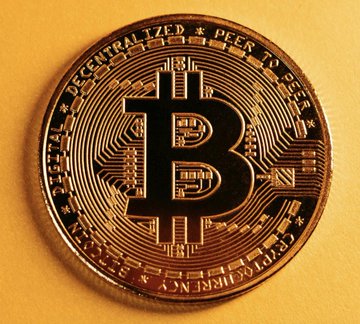 A close-up photograph of a physical Bitcoin token displaying the iconic "B" symbol and the inscription "DECENTRALIZED PEER TO PEER" on a gold-colored surface.