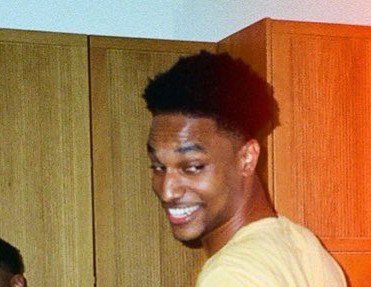 A close-up photo of a smiling young man with short dark hair, photographed in what appears to be an indoor setting with wooden cabinets visible in the background.