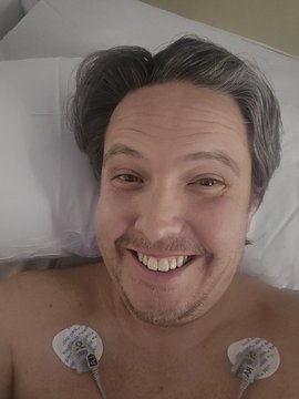 The image features a man lying down with two white earbuds attached to his ears, both of which are connected to an ECG machine. The man has a smile on his face as he looks directly at the camera. The background appears to be a hospital bed or a similar medical setting.