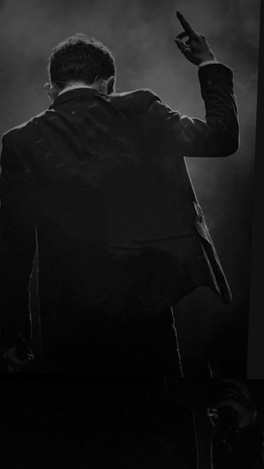 A black and white photograph of a man in a suit viewed from behind, pointing upward with his right hand against a smoky, atmospheric background.